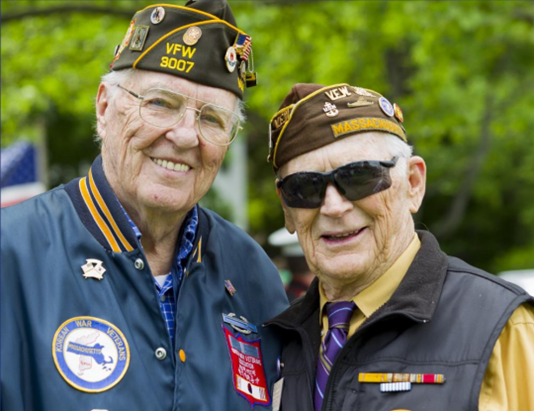 Two men wearing veteran caps smile outside, one with a patch-covered jacket and eyeglasses, and the other in sunglasses and a vest.